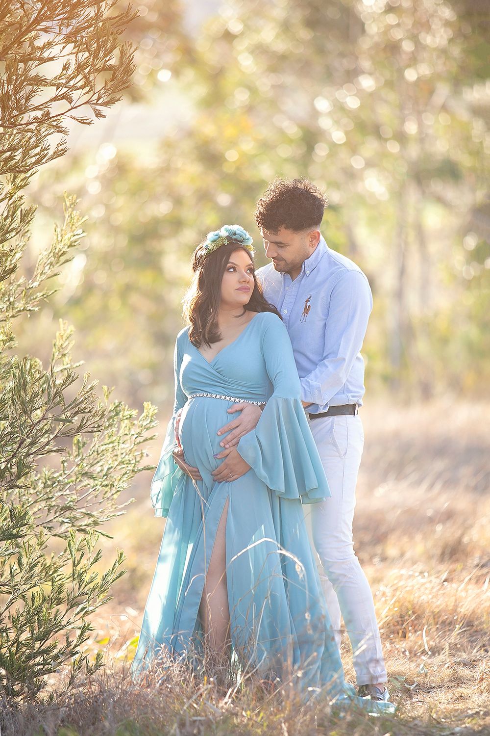 Pregnant woman in teal gown with floral crown embraced by partner in sunlit outdoor maternity portrait.
