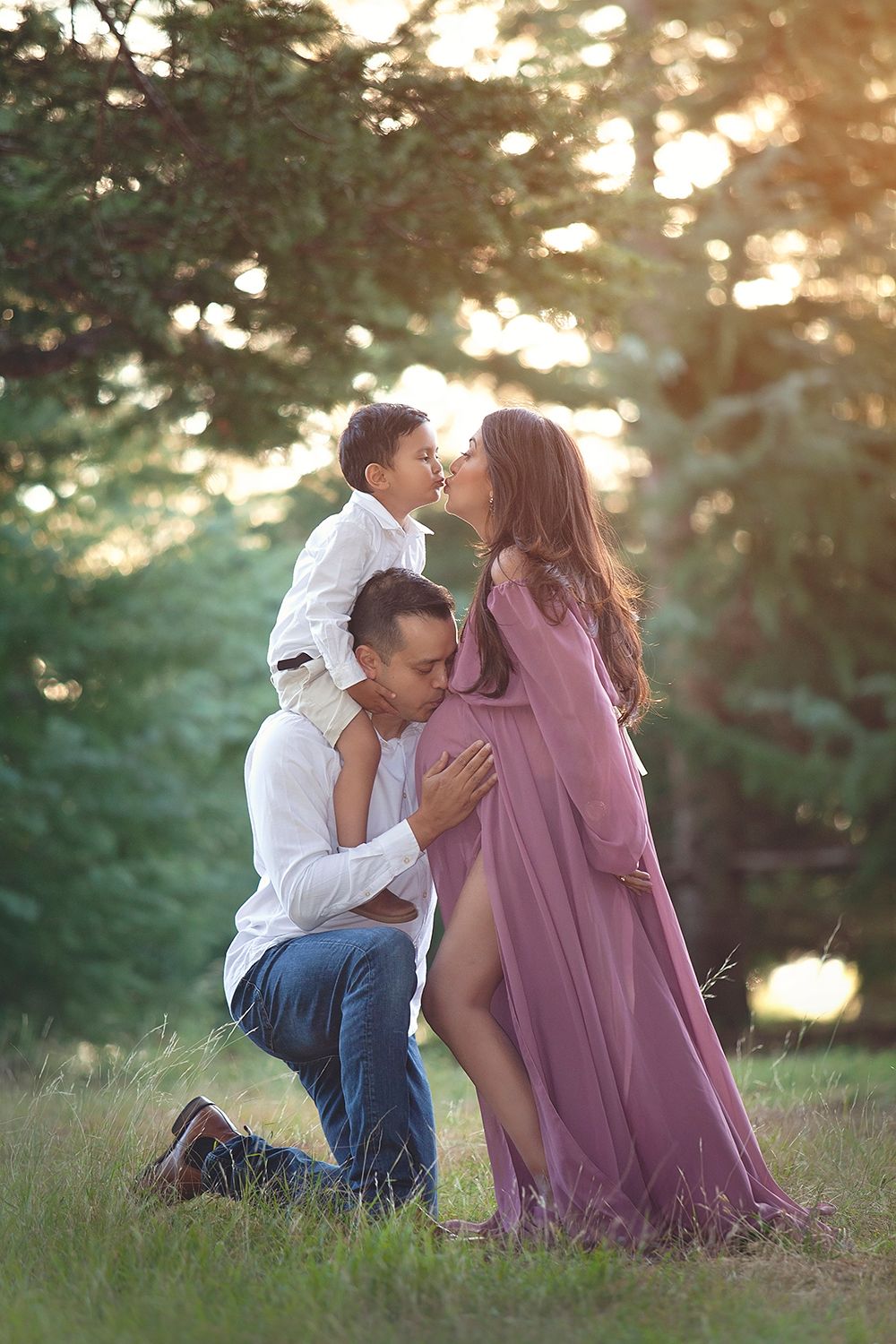 Family maternity photo with father kneeling, young son kissing pregnant mother in pink gown outdoors.