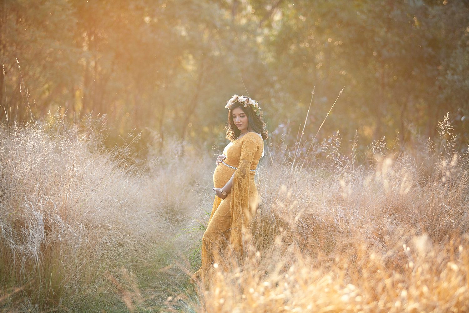 Pregnant woman in mustard yellow dress with floral crown standing in golden sunlit field at sunset.
