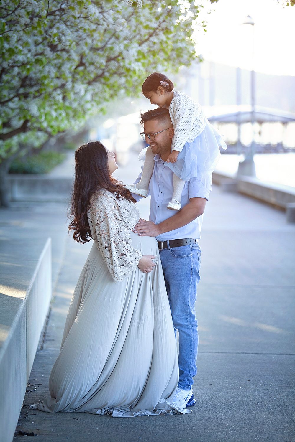Family maternity portrait with pregnant woman in grey gown, husband carrying child on shoulders outdoors.