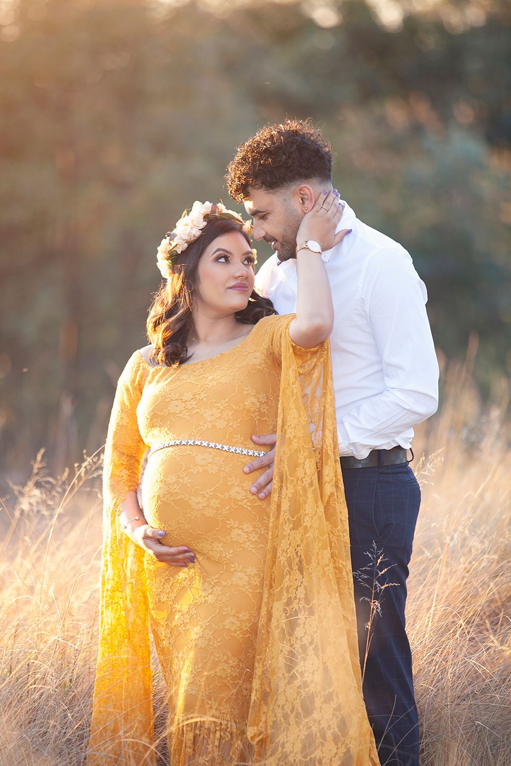 Couple sharing tender moment in golden field, pregnant woman in yellow lace gown during maternity shoot.