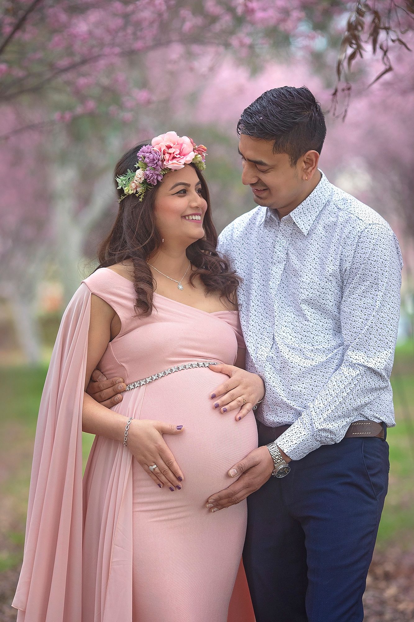 Expecting couple in pink cherry blossom garden, woman in pink maternity dress with flower crown.