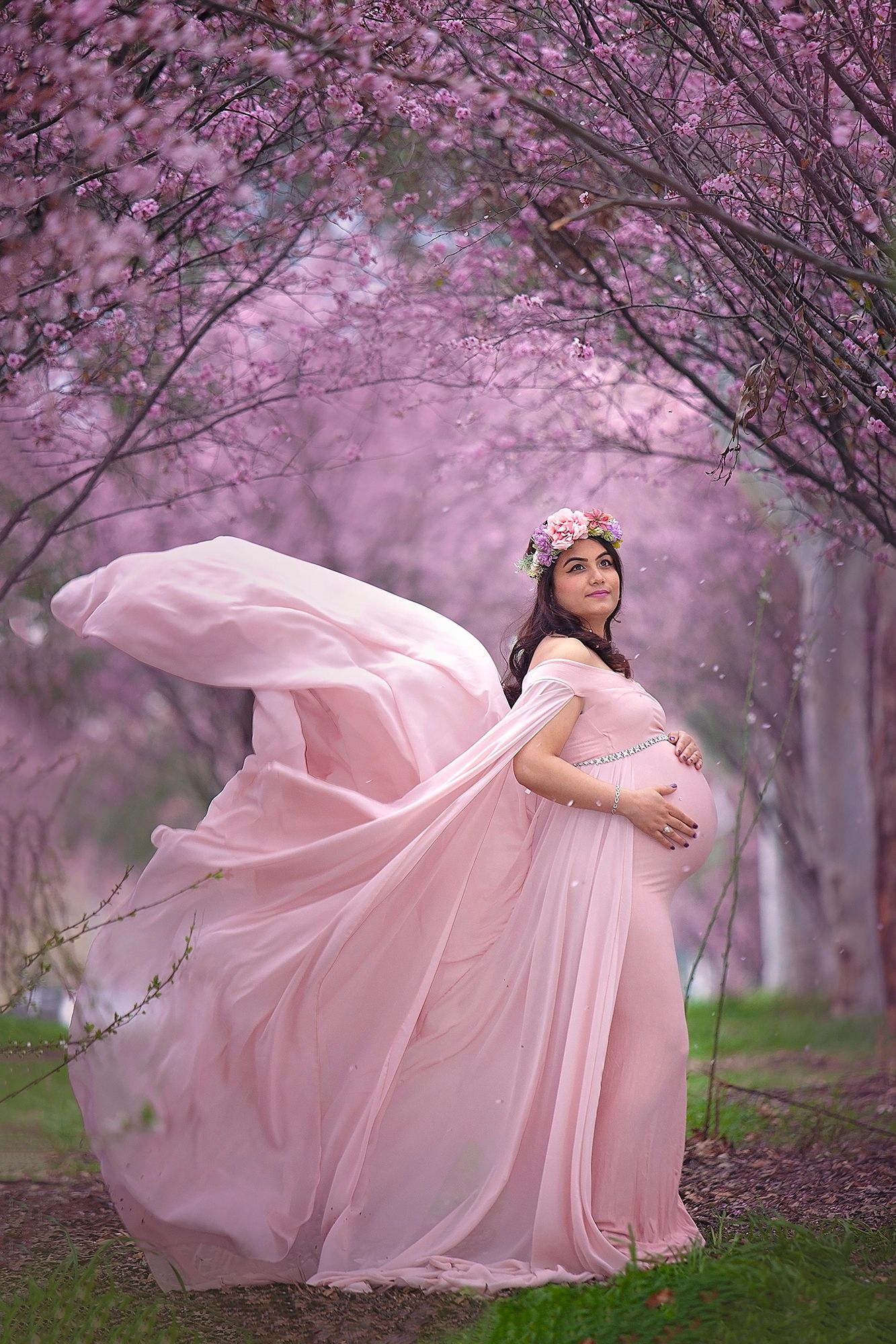 Pregnant woman in flowing pink gown with floral crown poses among blooming cherry blossom trees.