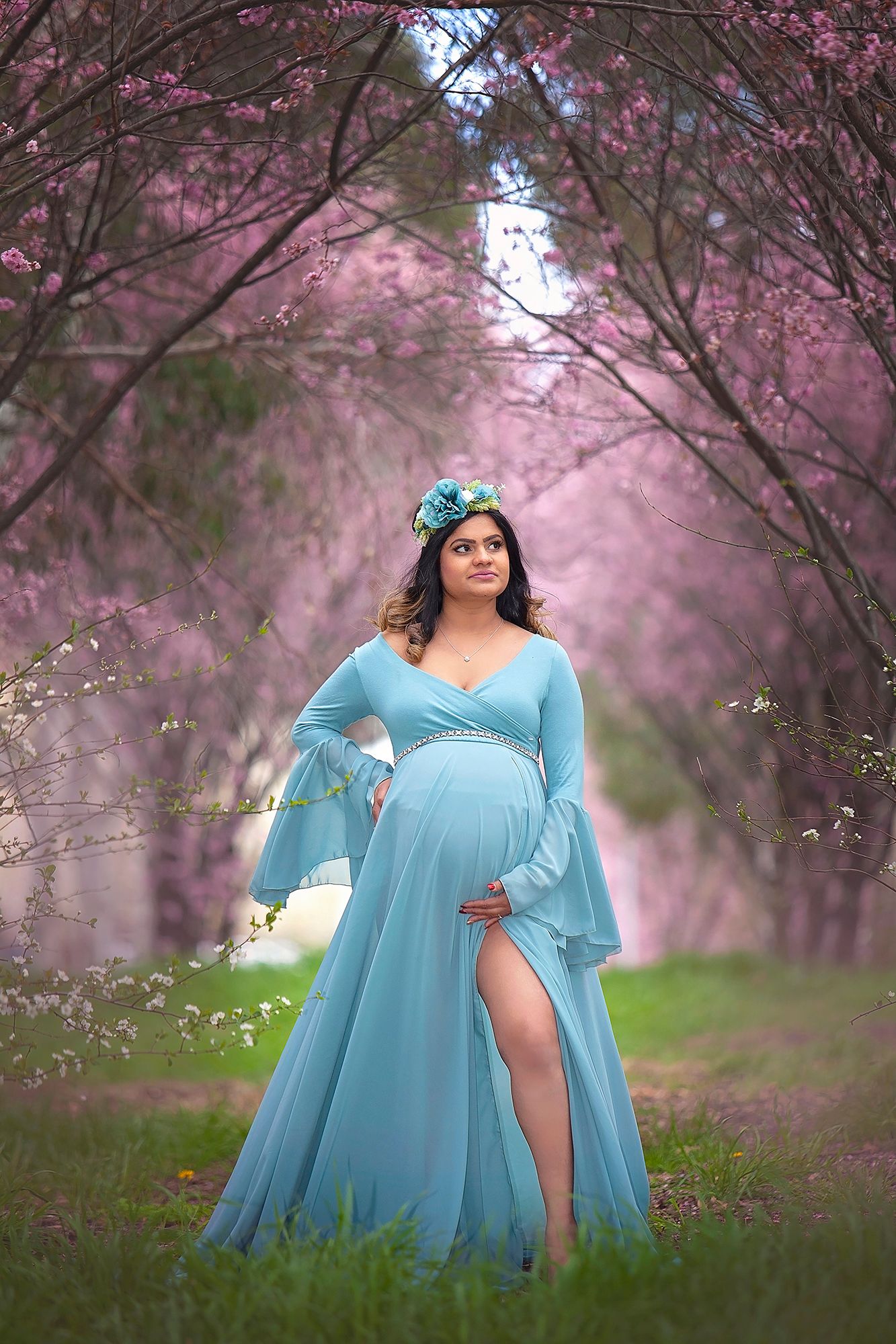 Pregnant woman in teal chiffon maternity gown with floral crown poses in cherry blossom garden.