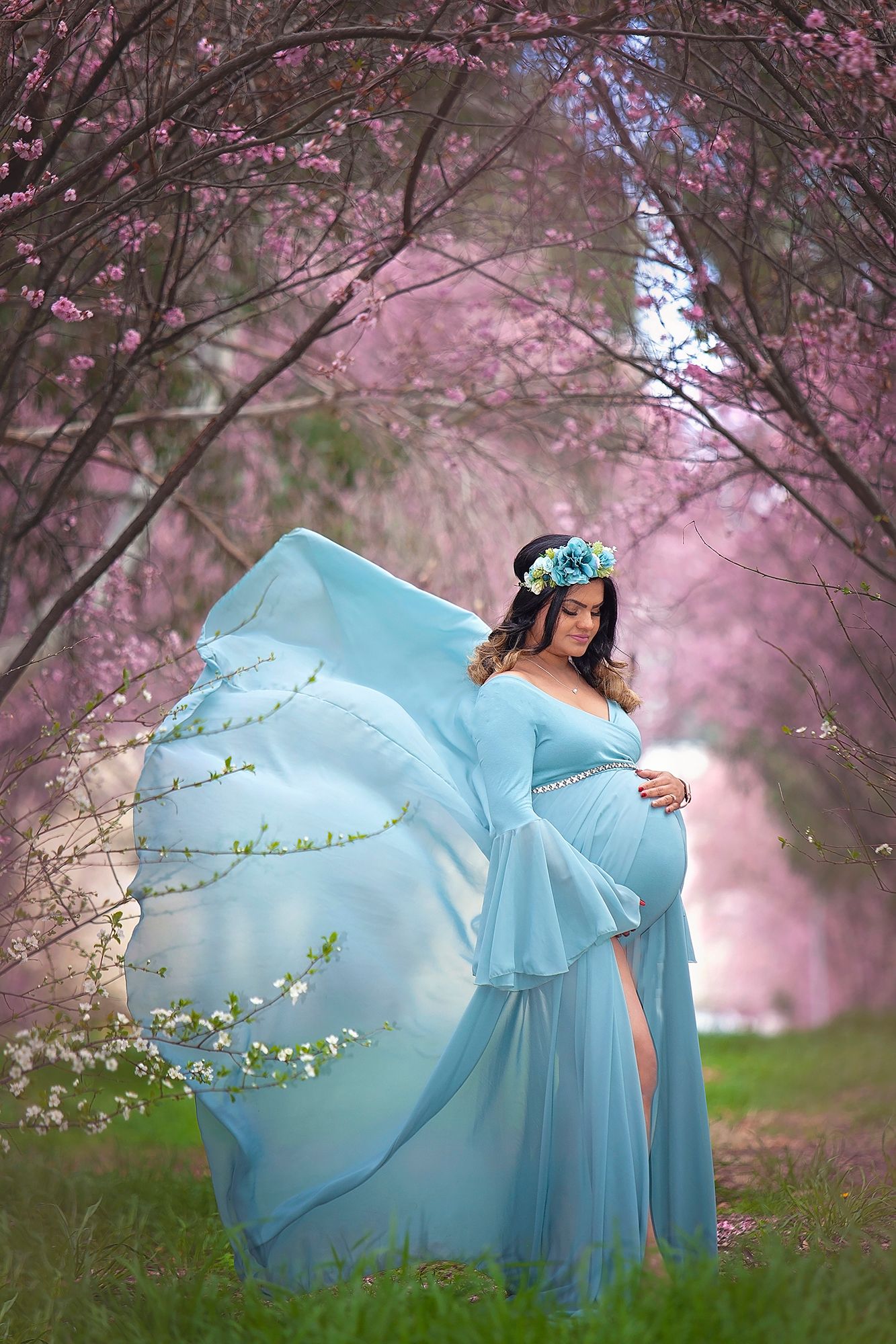 Pregnant woman in flowing teal gown with blue floral crown stands among pink cherry blossom trees.