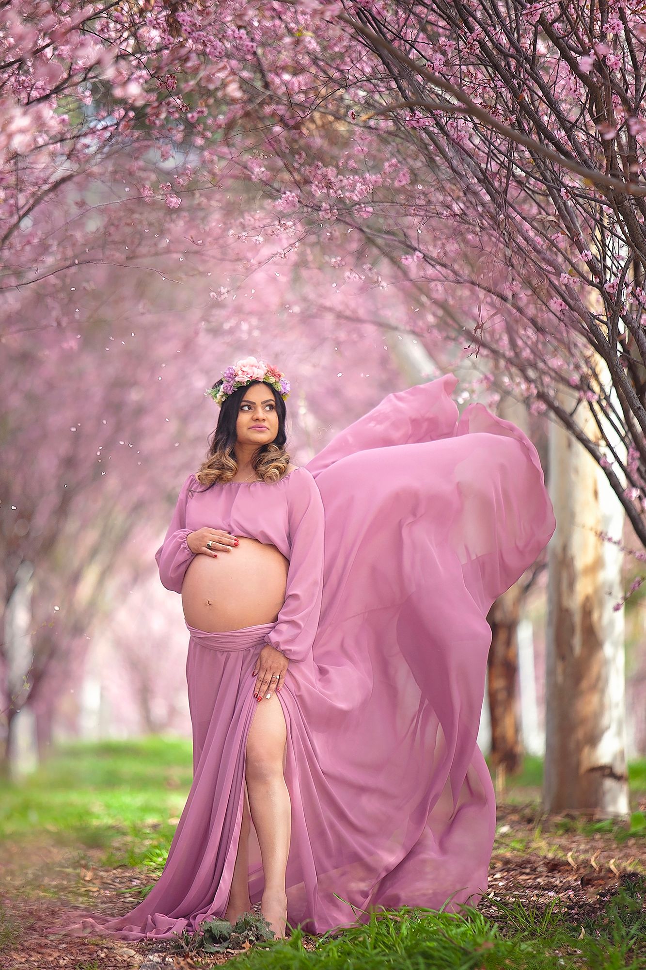 Pregnant woman in flowing mauve gown with floral crown cradles belly among pink cherry blossoms.