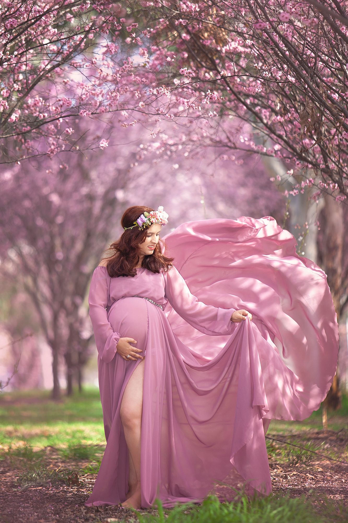 Pregnant woman in billowing pink mauve gown with floral crown poses in cherry blossom garden.