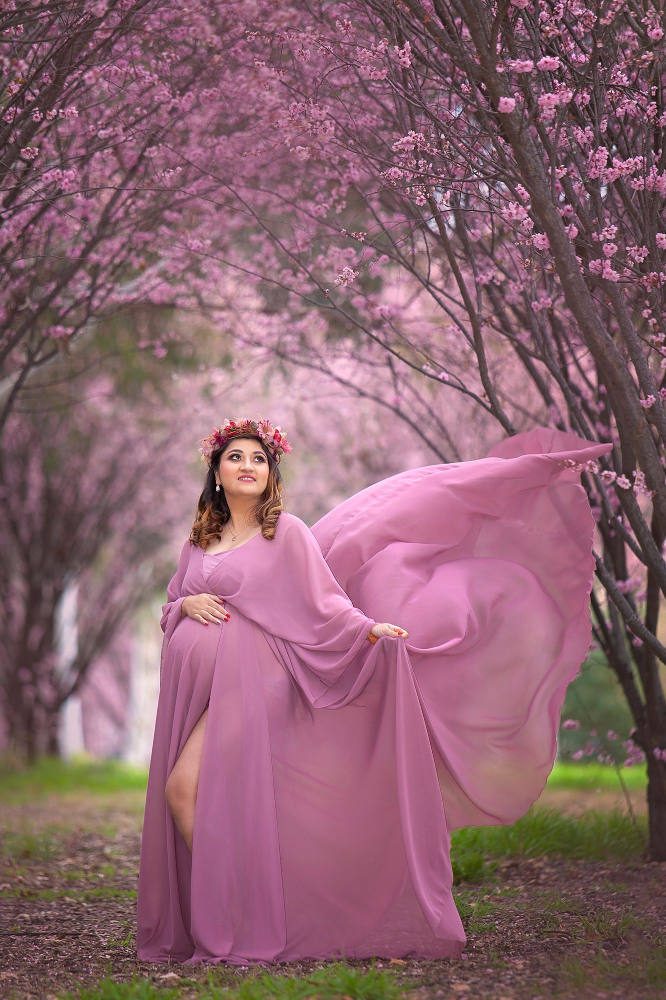 Pregnant woman in flowing pink chiffon gown with red floral crown poses in cherry blossom garden.