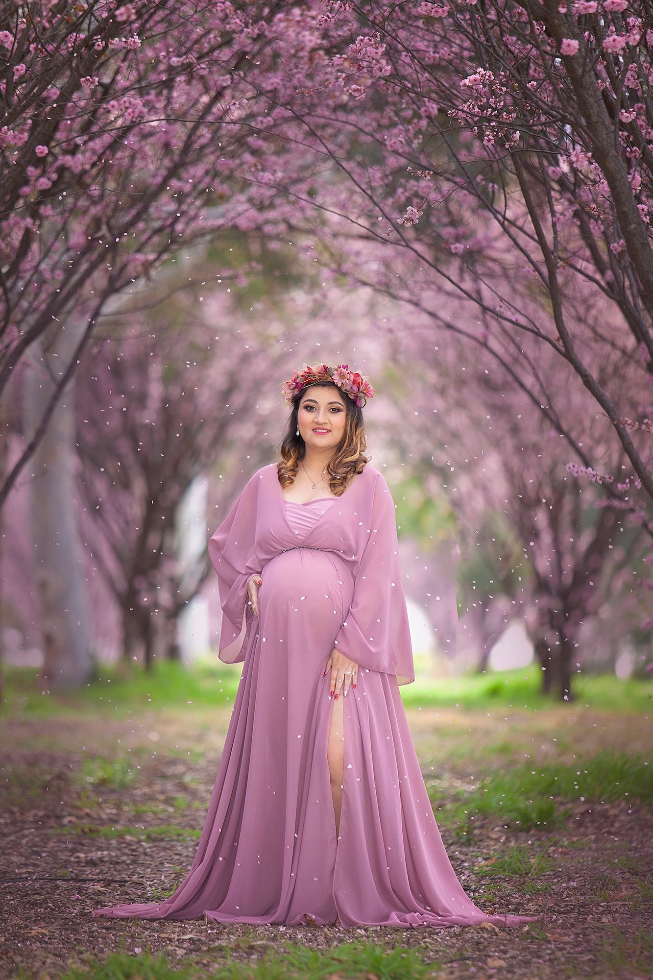Pregnant woman in sheer mauve gown with pink floral crown stands in blooming cherry blossom garden.