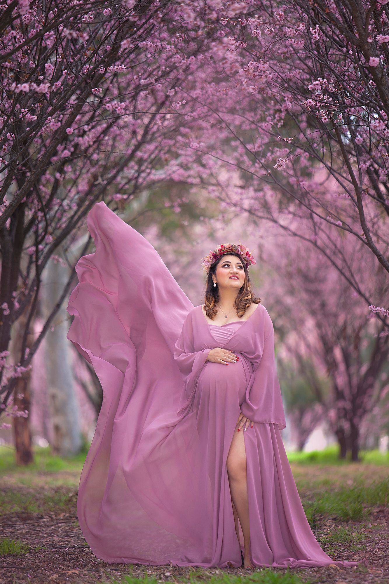 Pregnant woman in flowing pink gown with floral crown poses among blooming pink cherry blossom trees.