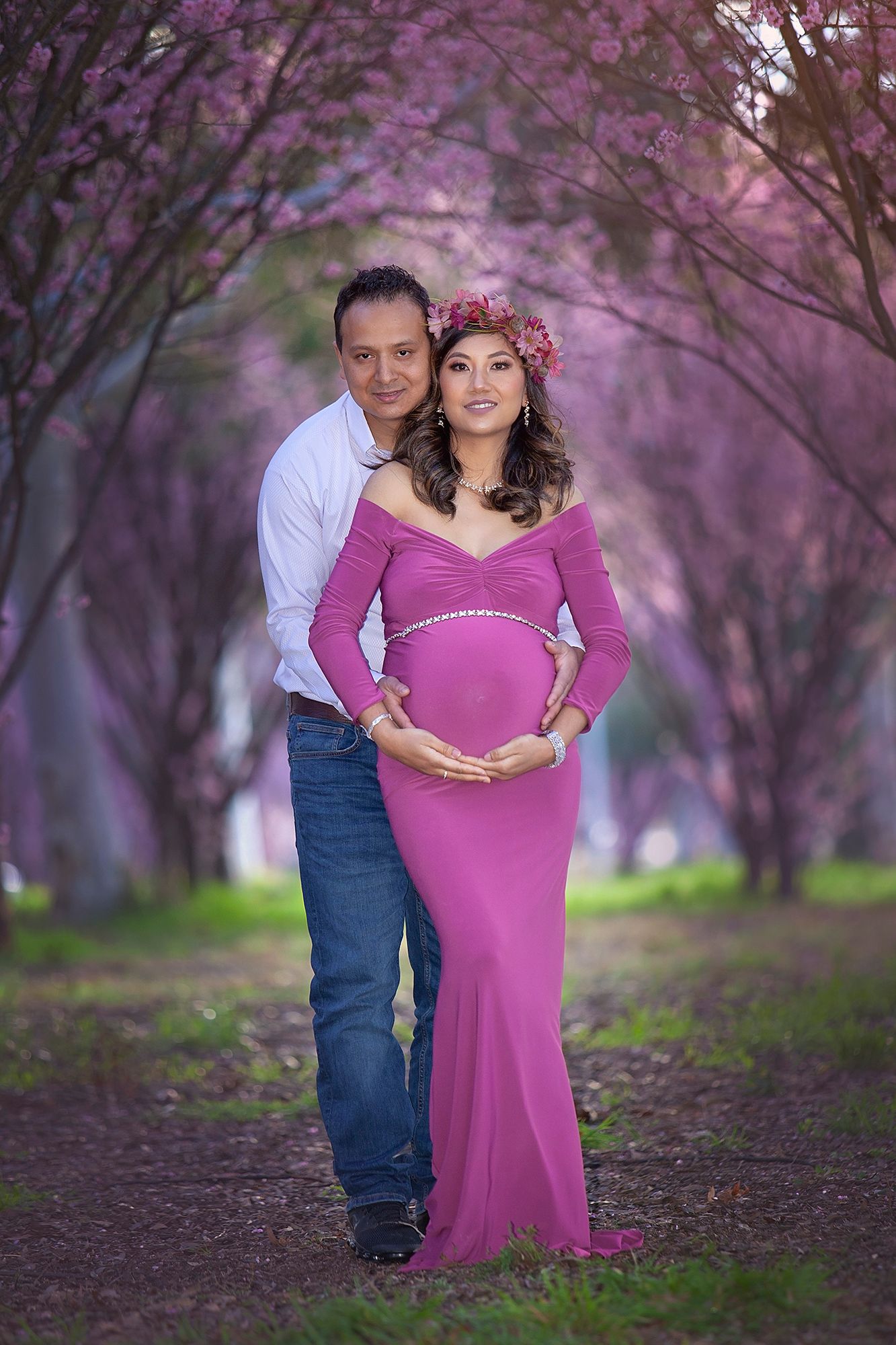 Couple poses in blooming pink tree tunnel, woman in fitted pink maternity gown with floral crown.