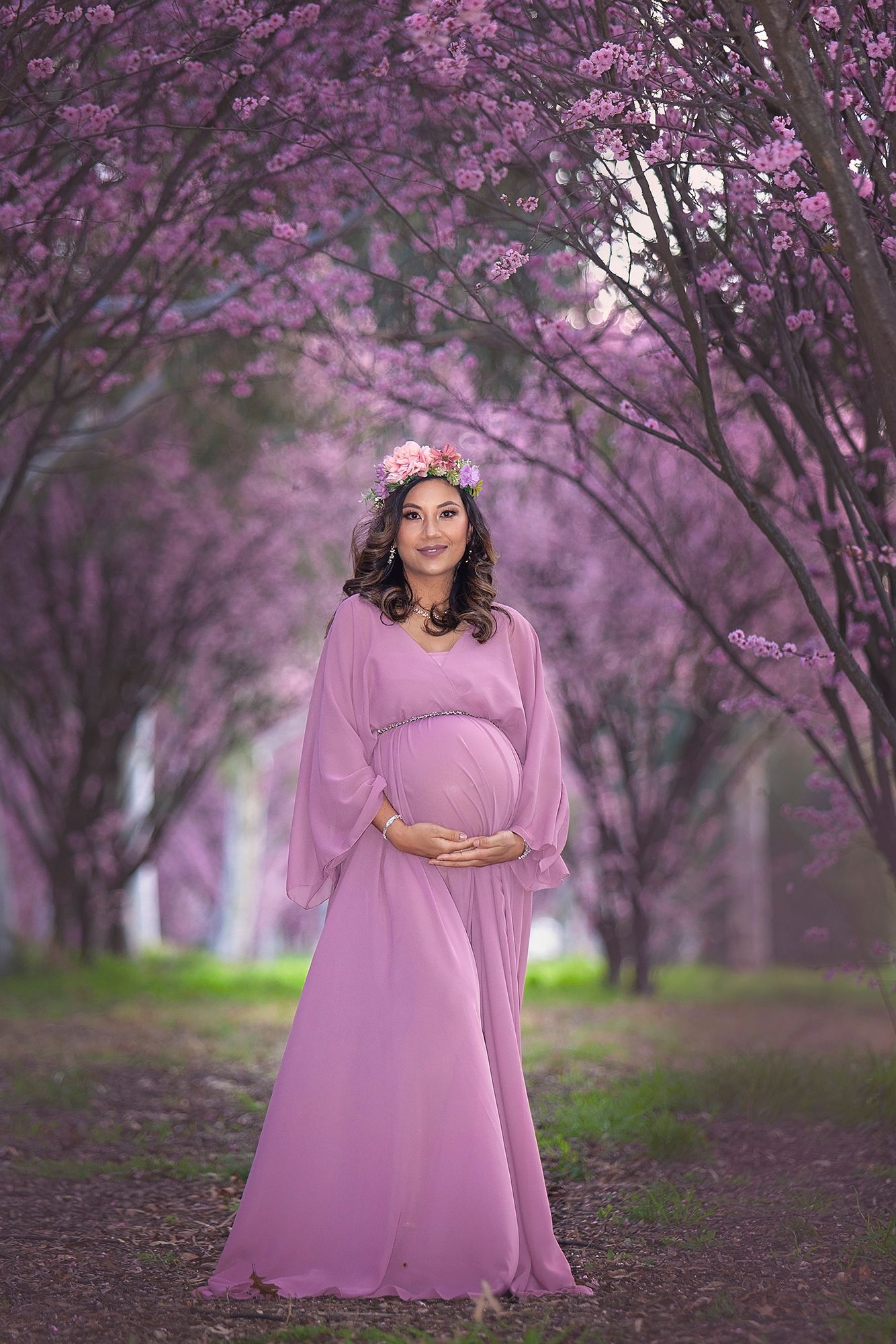 Pregnant woman in purple chiffon gown and floral crown smiles in a pink cherry blossom garden.