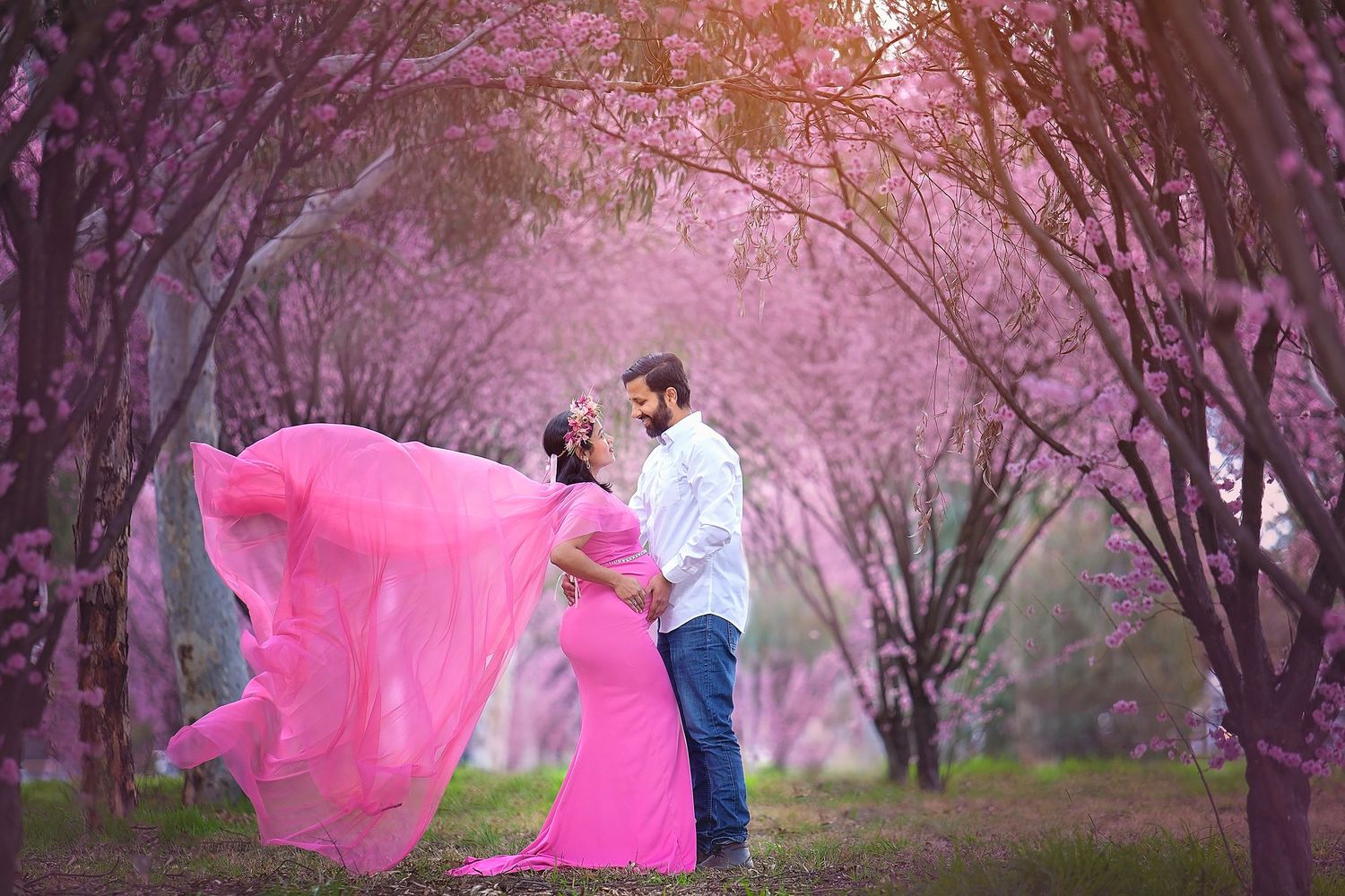 Couple shares a romantic moment in pink flowing maternity gown among blooming cherry blossom trees.