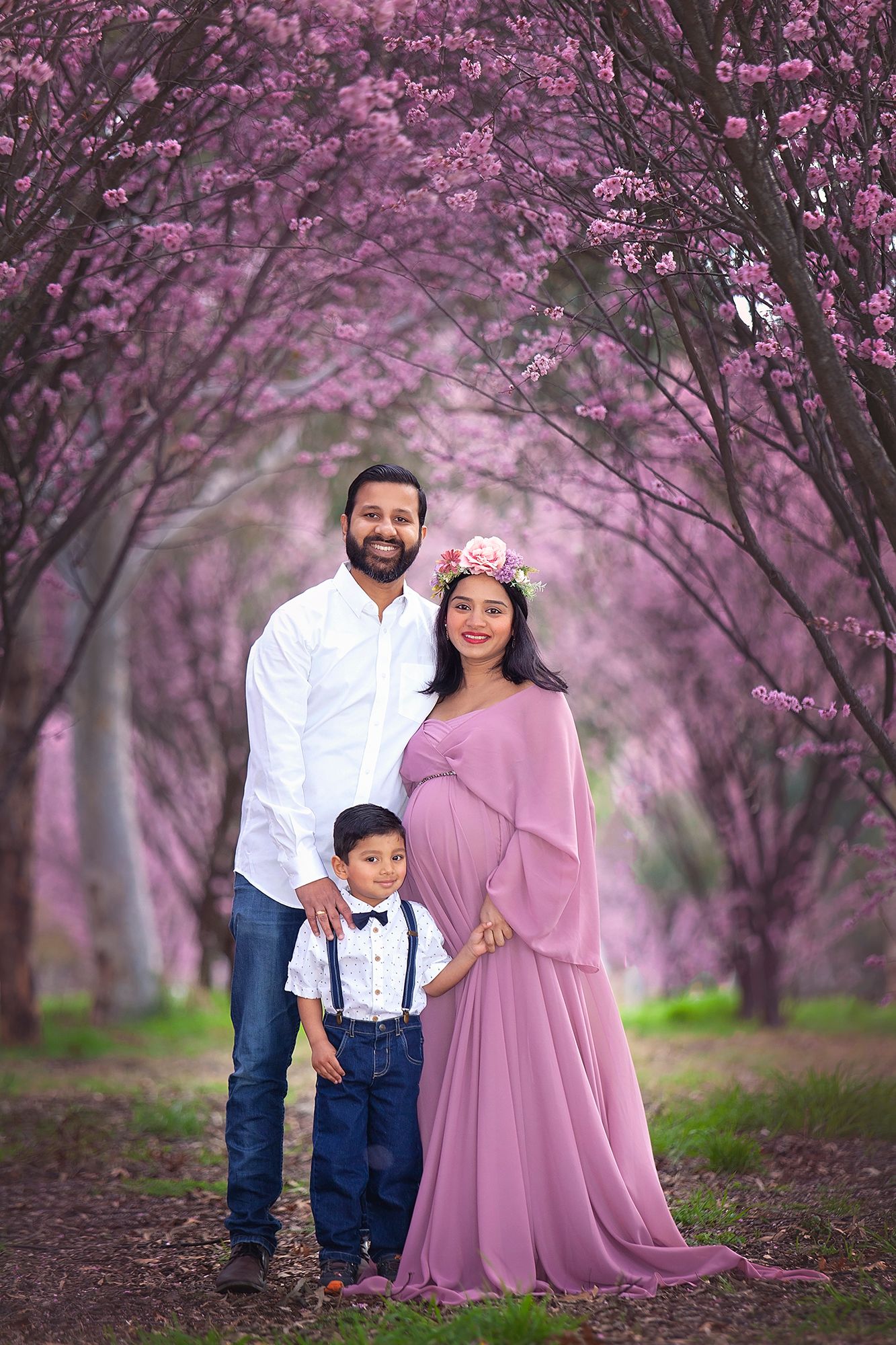 Family maternity photo with pregnant woman in pink gown, husband, and young son in cherry blossoms.