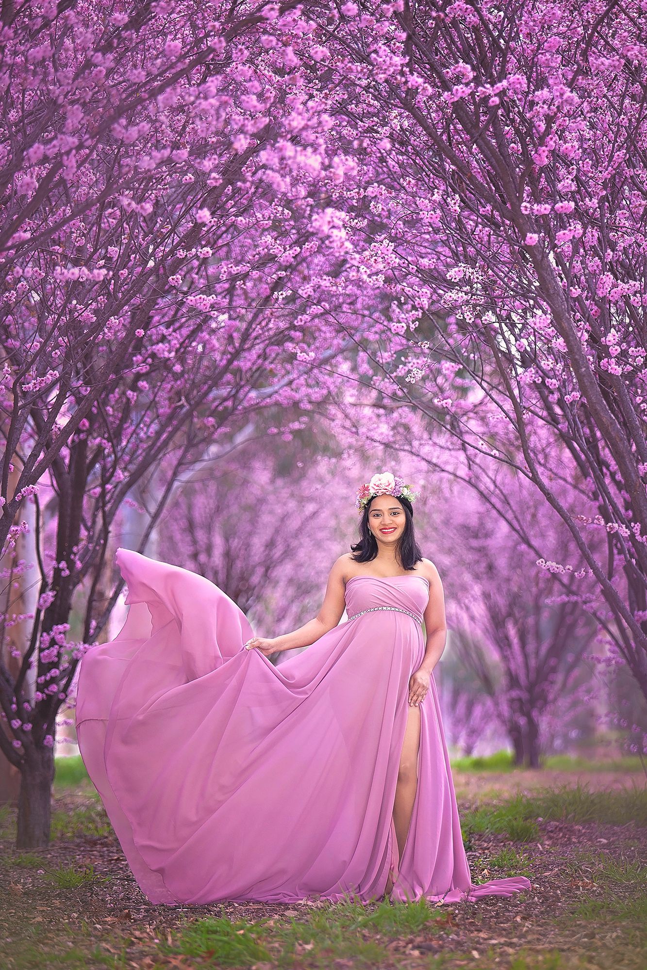 Pregnant woman in flowing pink gown with white floral crown poses in a pink cherry blossom tunnel.