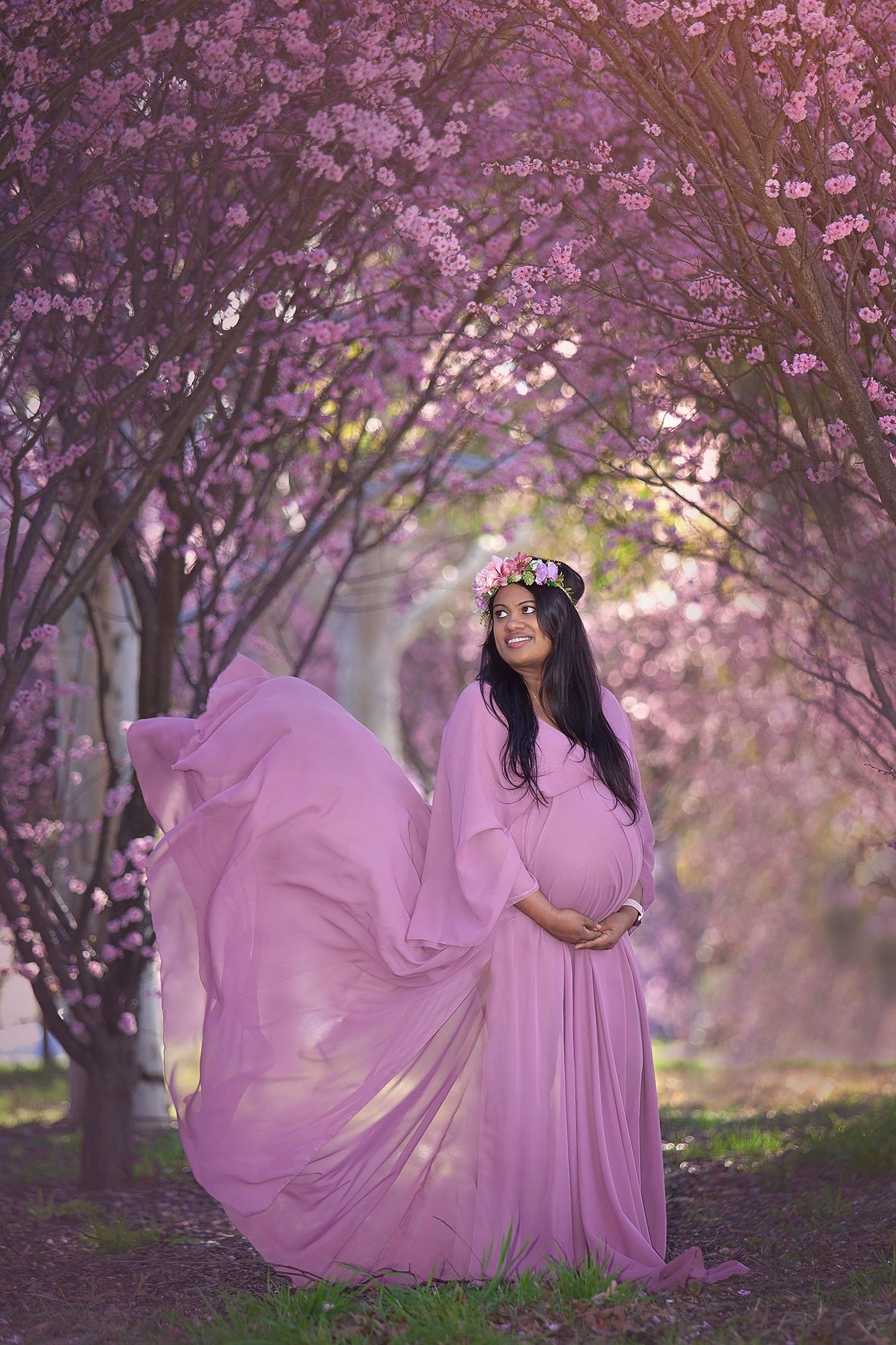 Smiling pregnant woman in flowing pink gown and floral crown poses in pink cherry blossom garden.