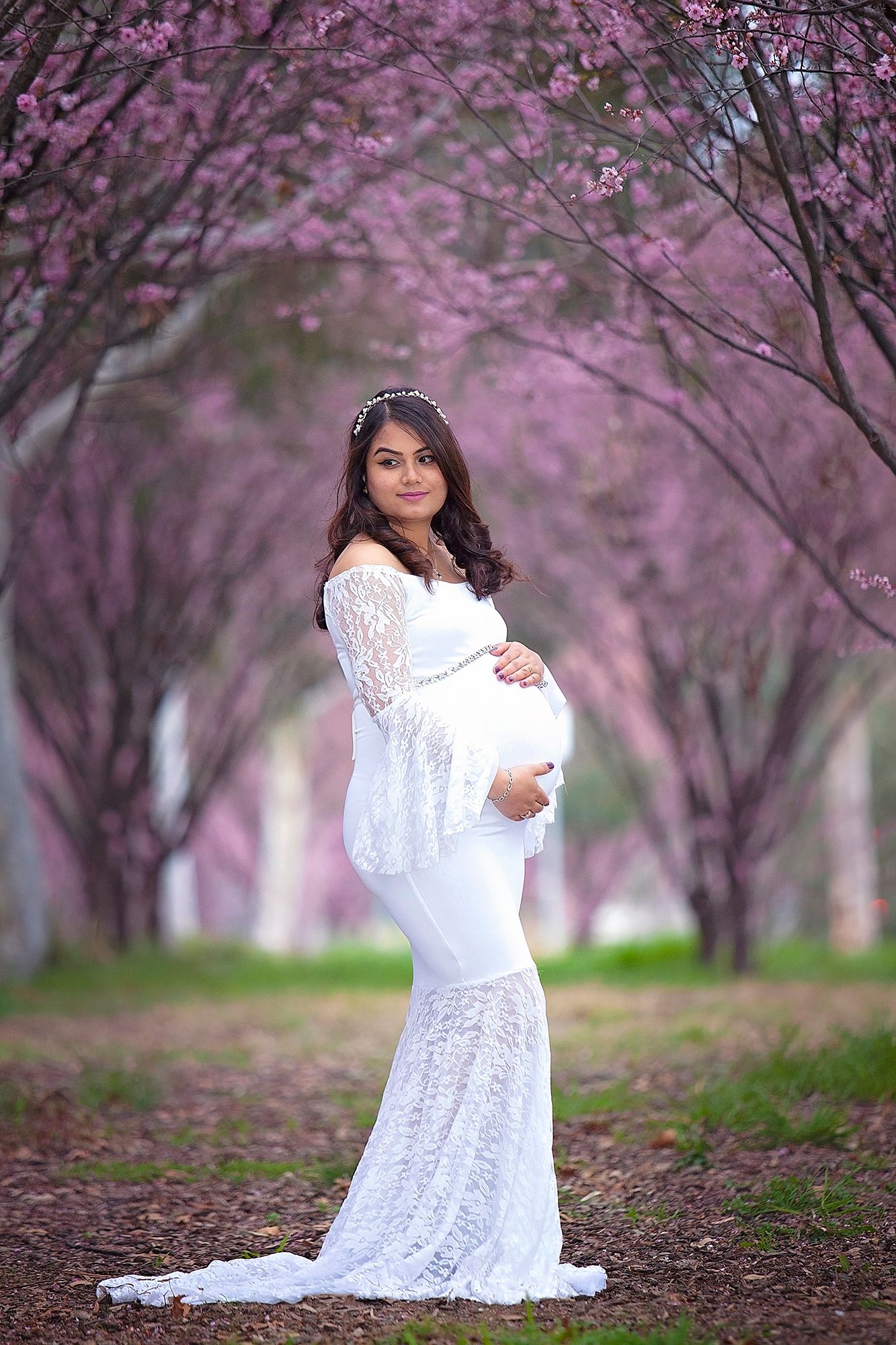 Pregnant woman in elegant white lace maternity gown poses in a pink cherry blossom tree tunnel.