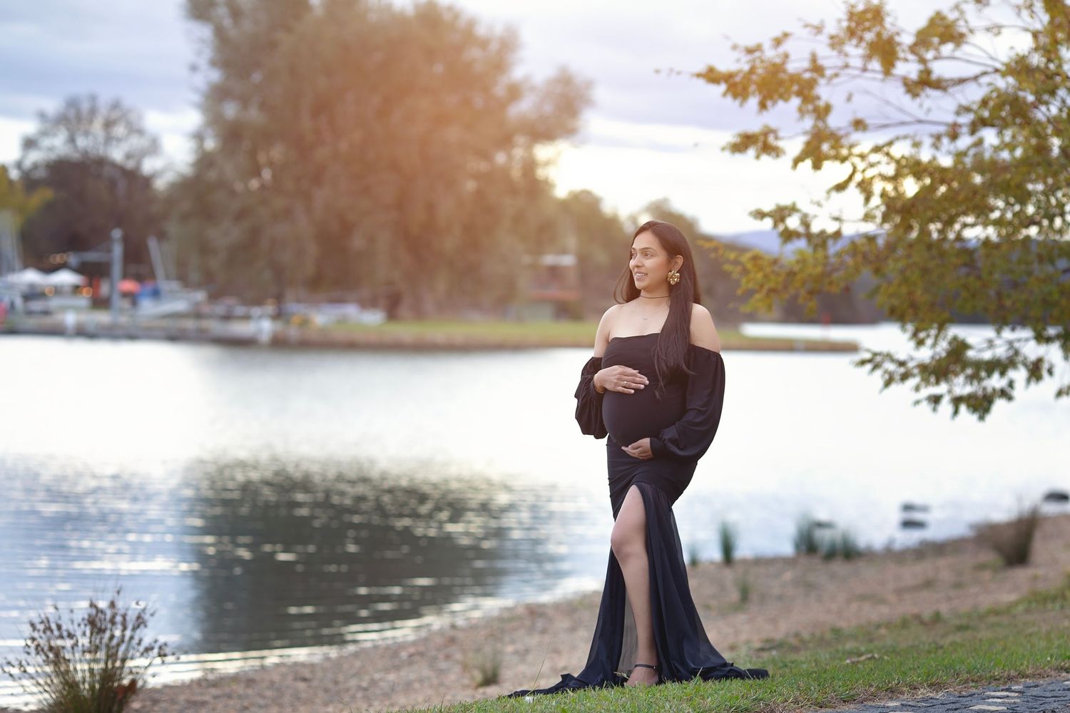 Pregnant woman in elegant black gown posing by a sunlit lake during golden hour.