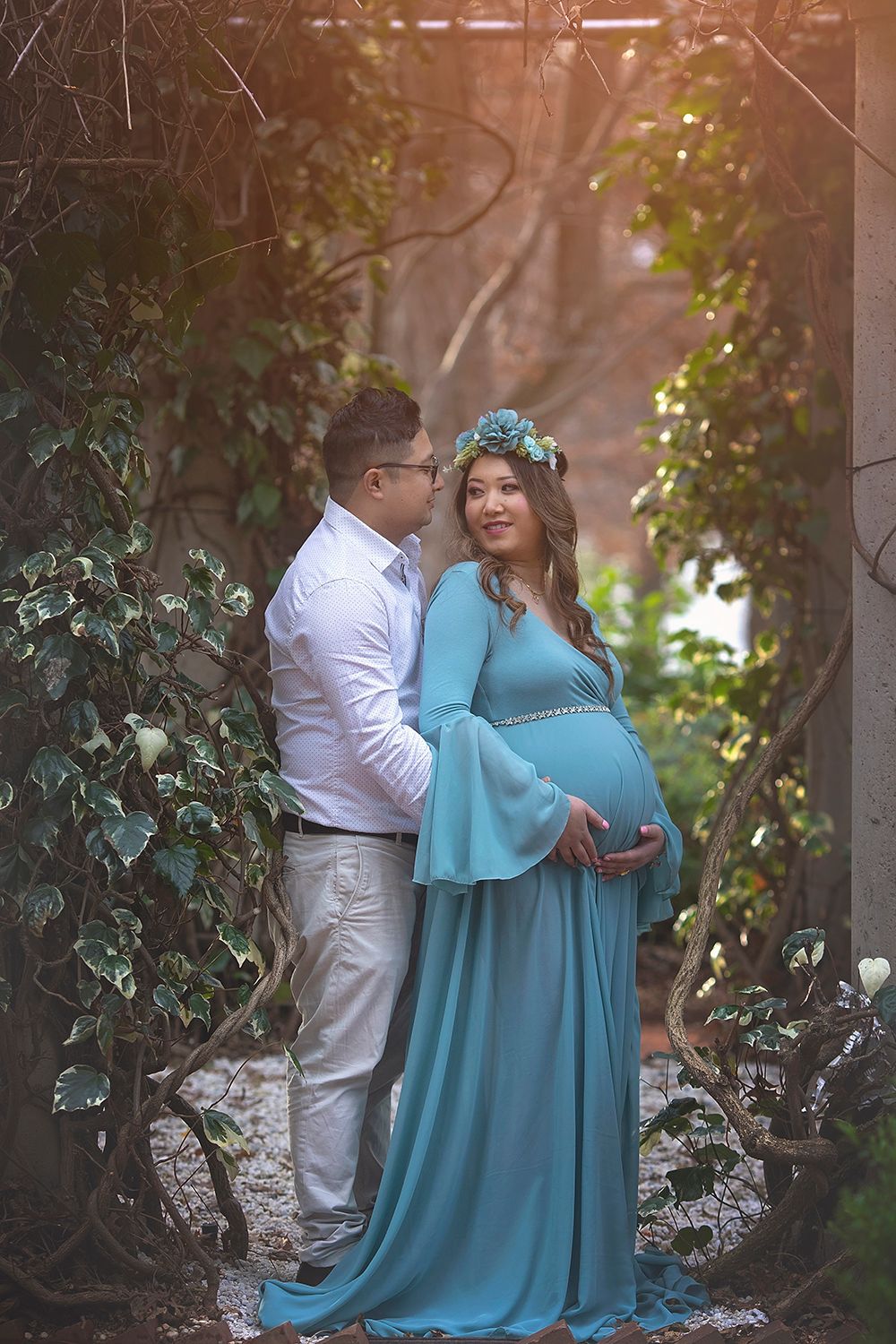 Pregnant couple embracing tenderly in a sunlit garden archway surrounded by lush green ivy.