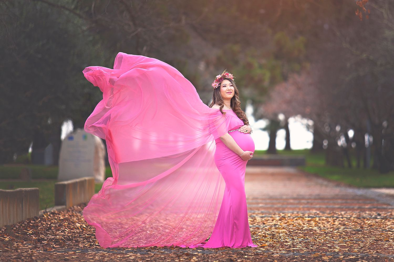 Pregnant woman in a flowing pink gown with fabric billowing in the wind in an autumn park.