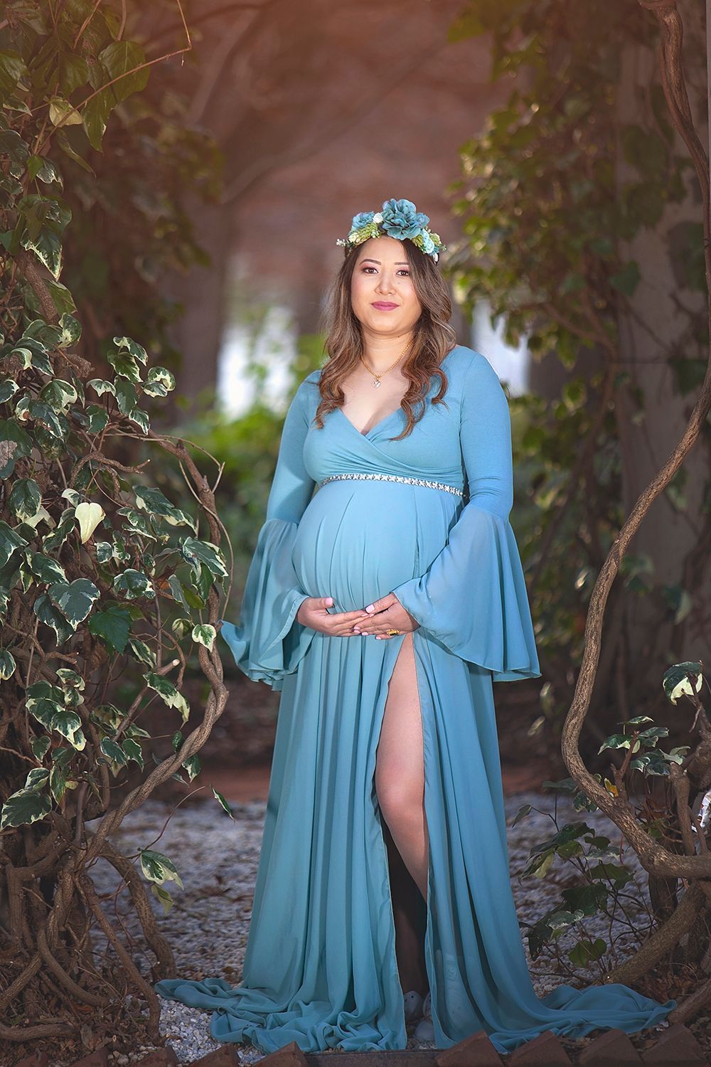 Pregnant woman in a teal gown with a floral crown standing gracefully among twisted vines outdoors.