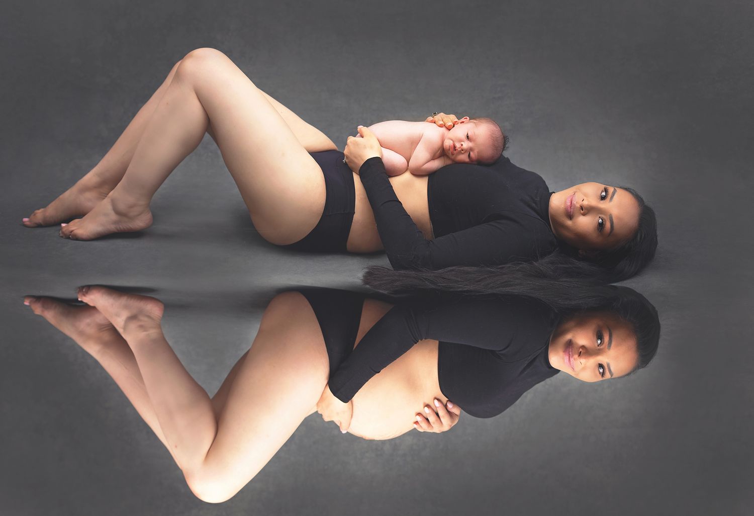 Pregnant woman in black bodysuit lying with newborn baby on reflective surface.