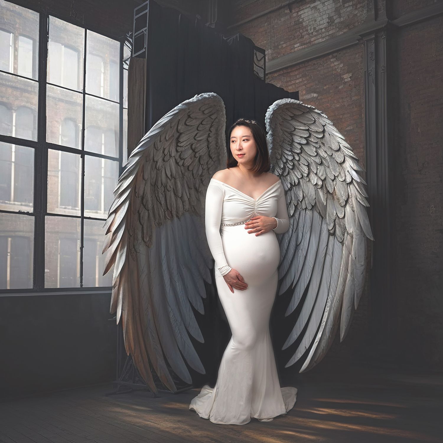 Pregnant woman in white dress stands before large angel wings in industrial loft maternity photography session.