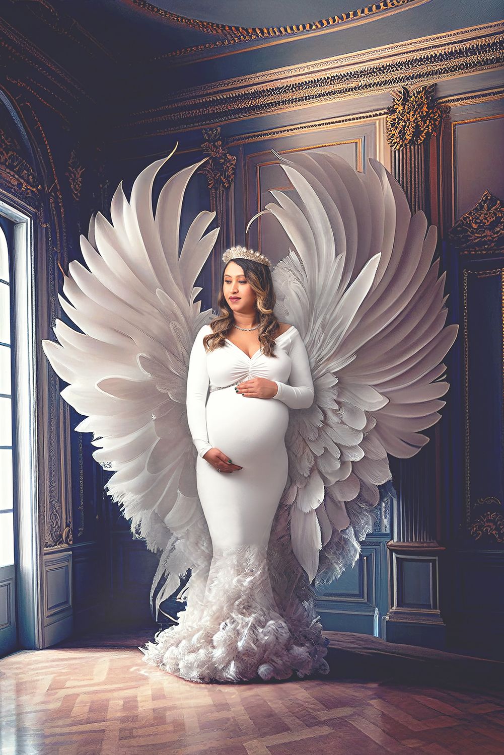 Pregnant woman wearing crown and white gown stands before large angel wings in ornate room for maternity portrait.