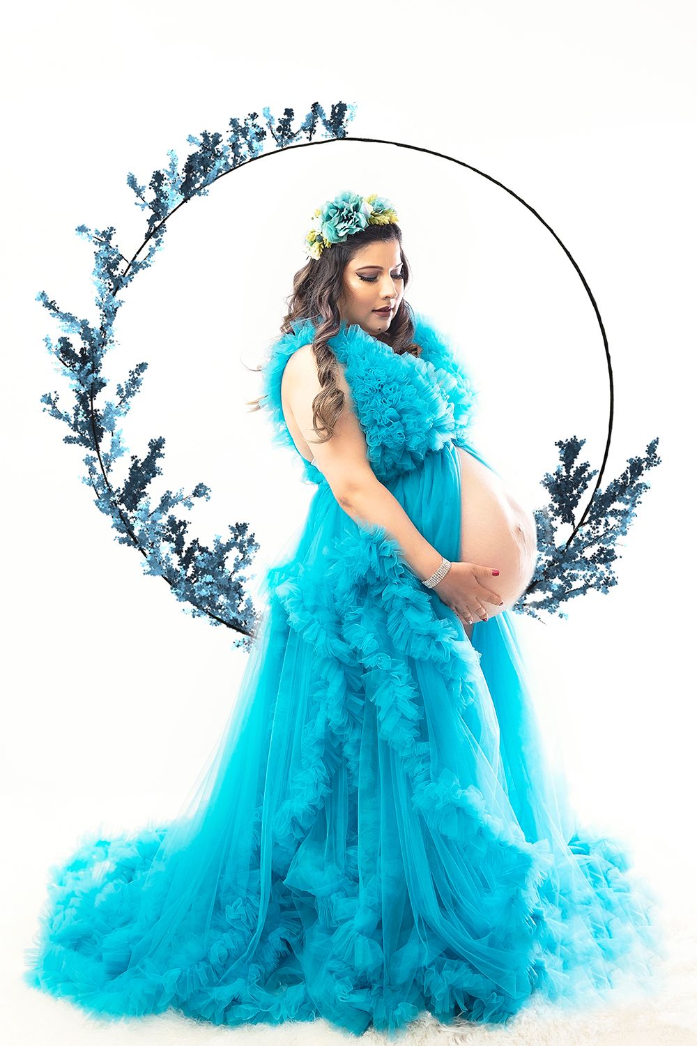 Pregnant woman in flowing turquoise tulle gown with floral crown poses in circular floral frame for maternity shoot.