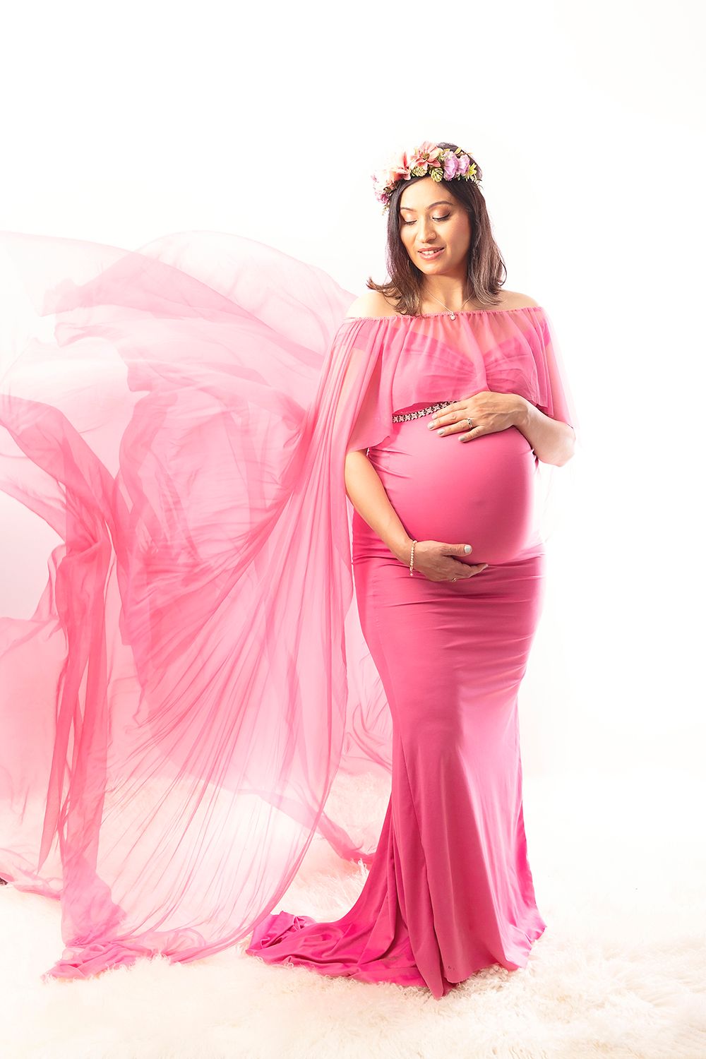 Pregnant woman in pink off-shoulder gown with floral crown poses with flowing fabric for elegant maternity portrait.