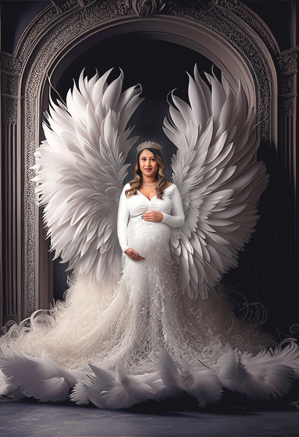 Pregnant woman in white lace gown with large angel wings and tiara poses in elegant archway.