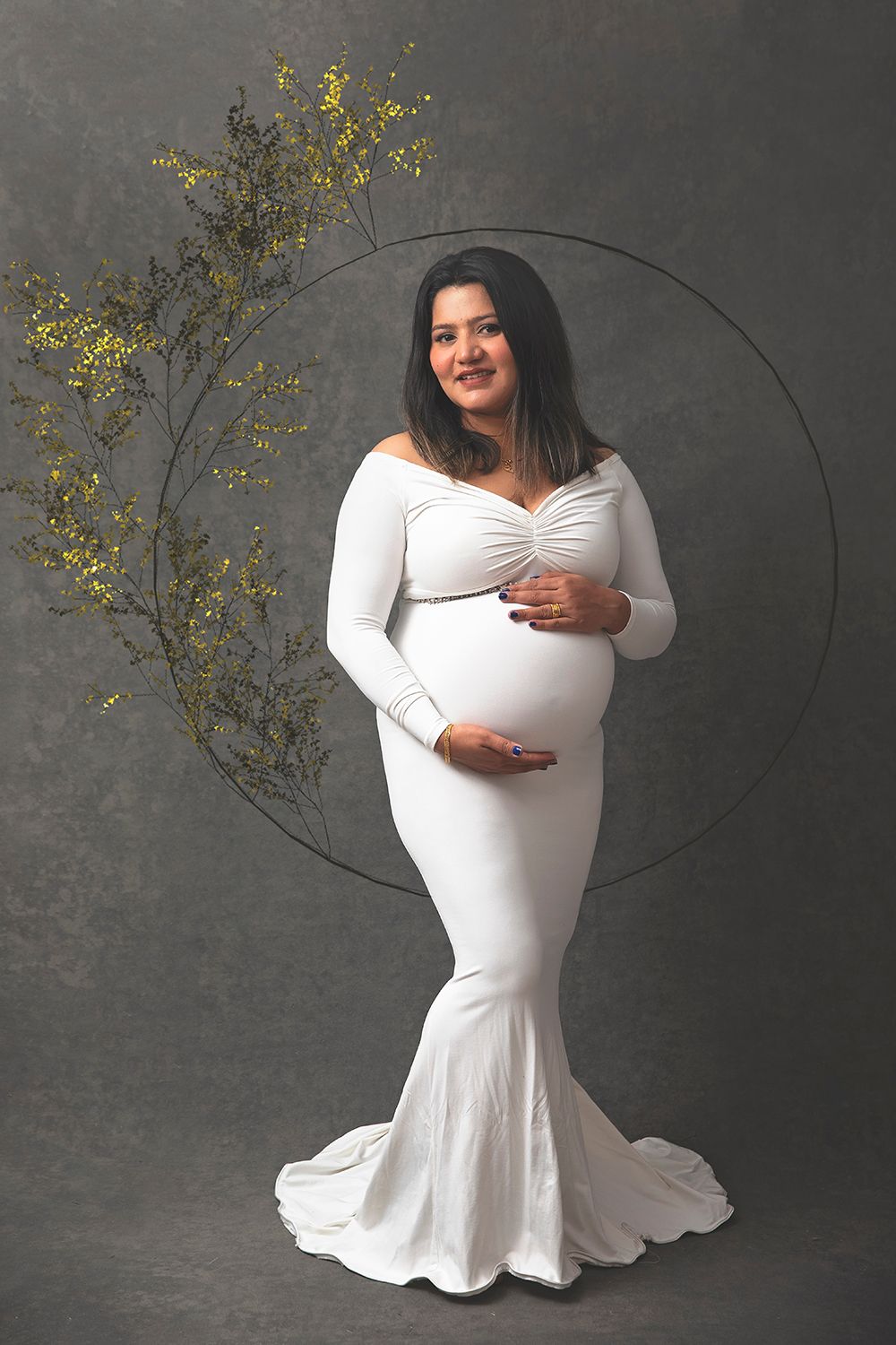 Pregnant woman in white mermaid gown poses before circular floral backdrop on gray background.