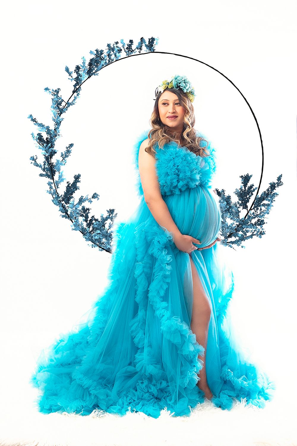 Pregnant woman in turquoise ruffled gown with floral crown poses in circular blue floral wreath.