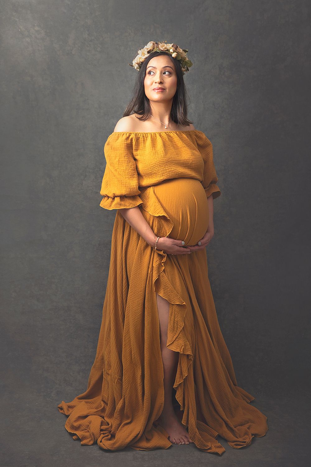 Pregnant woman in mustard off-shoulder gown with flower crown poses on gray studio background.