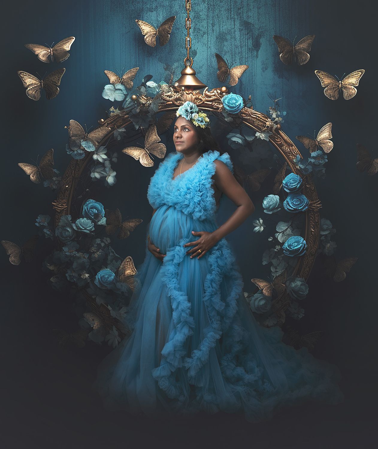 Pregnant woman in blue ruffled gown surrounded by butterflies and roses in dramatic blue studio.
