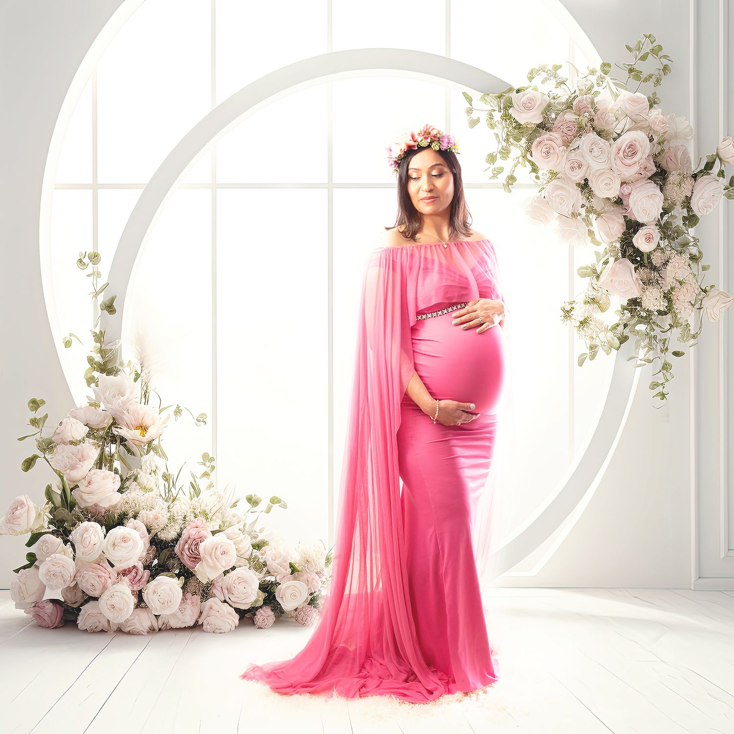 Pregnant woman in pink tulle gown with floral crown poses before white arch with roses.