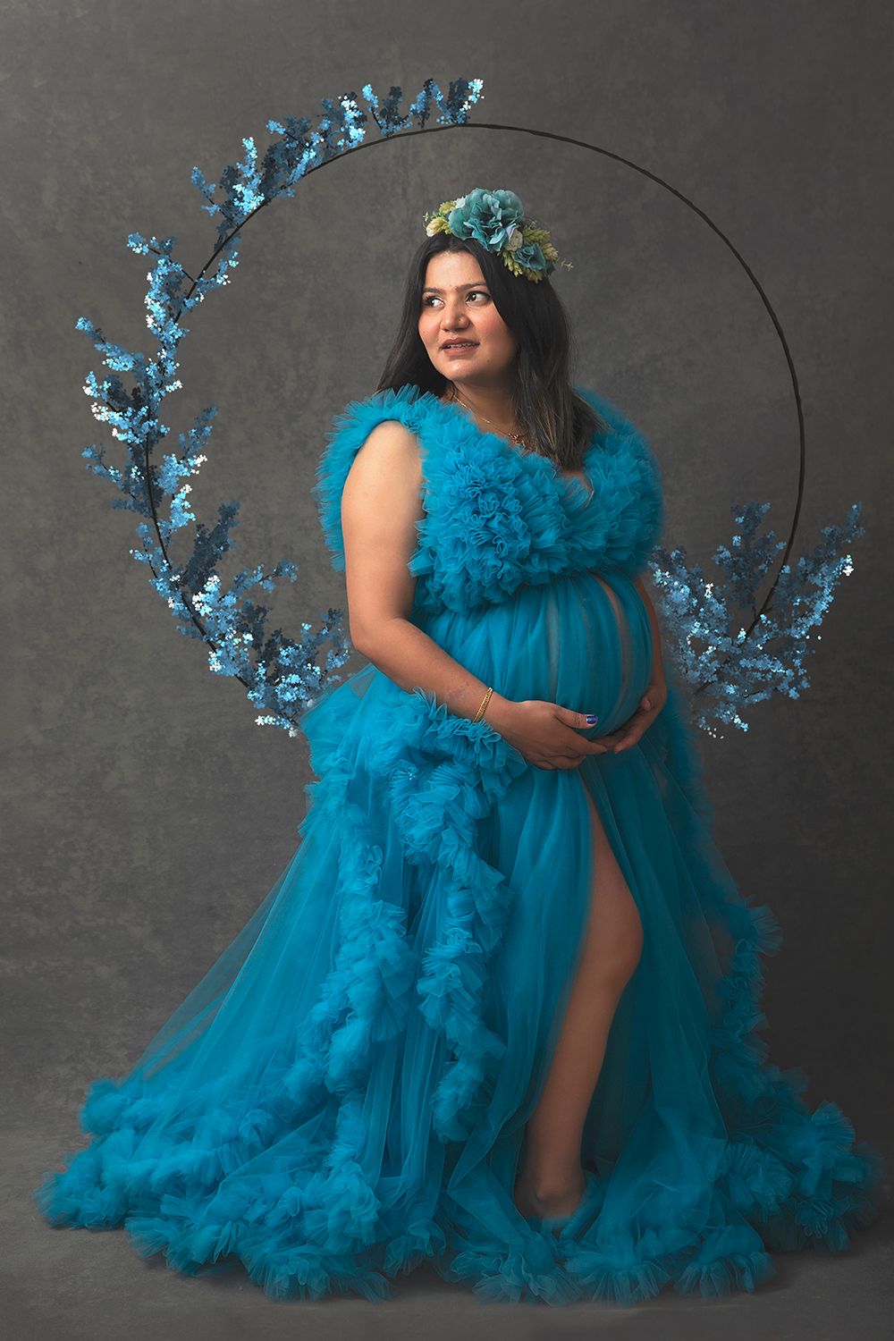 Pregnant woman in teal ruffled gown with floral crown poses before circular floral arch.