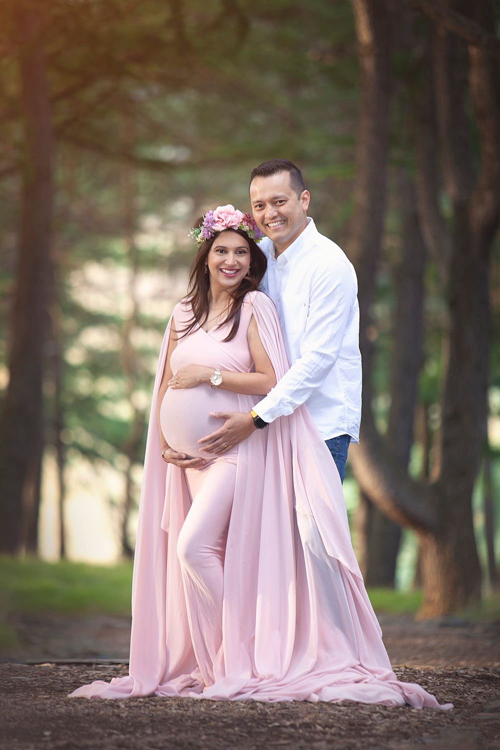 Smiling couple in forest, pregnant woman in pink gown with floral crown embraced by partner.
