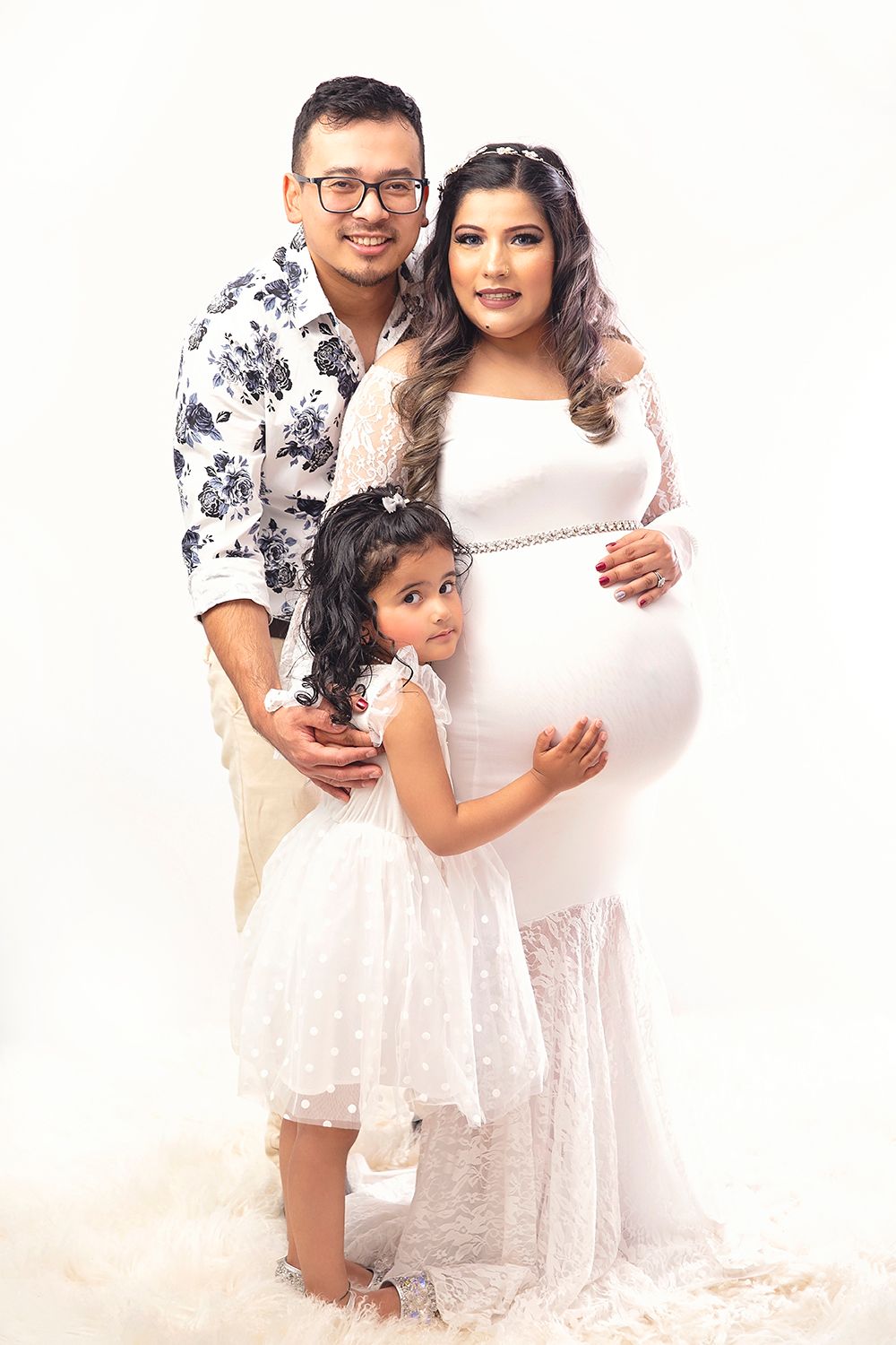 Family maternity photo with pregnant woman in white gown, husband, and young daughter.