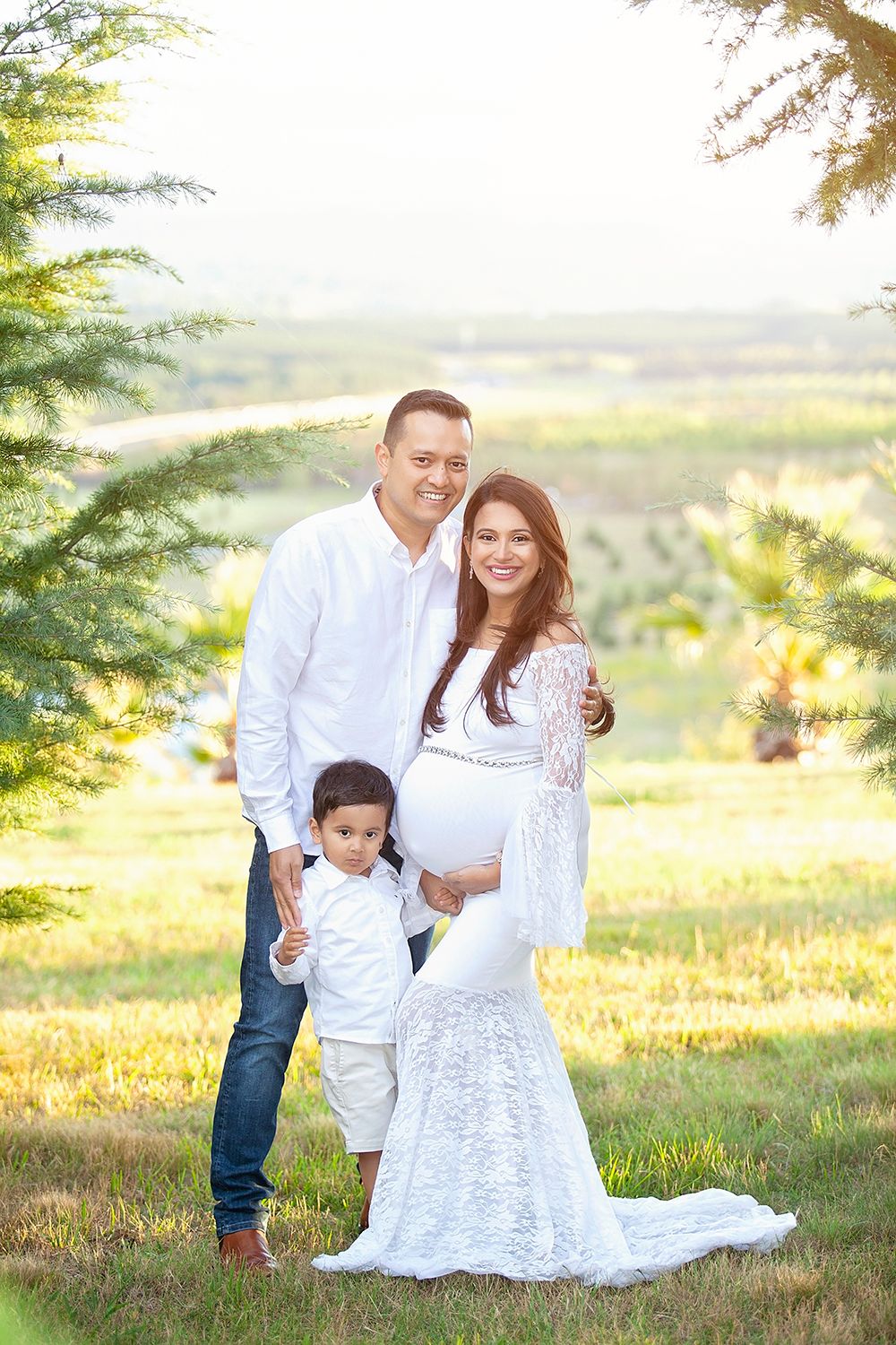 Family maternity portrait with pregnant woman in white lace gown, husband and son in sunny park.