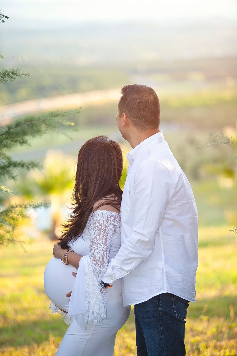 Couple maternity photo from behind, pregnant woman in white lace dress embraced by partner outdoors.