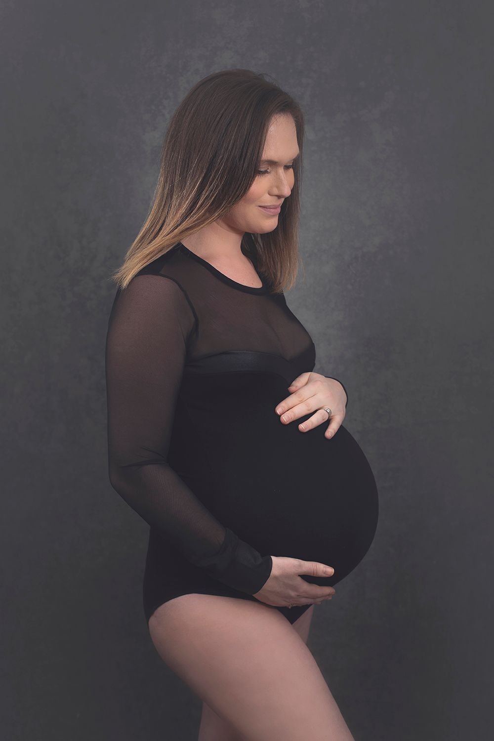 Pregnant woman in black mesh bodysuit cradling her baby bump against a dark gray studio background.