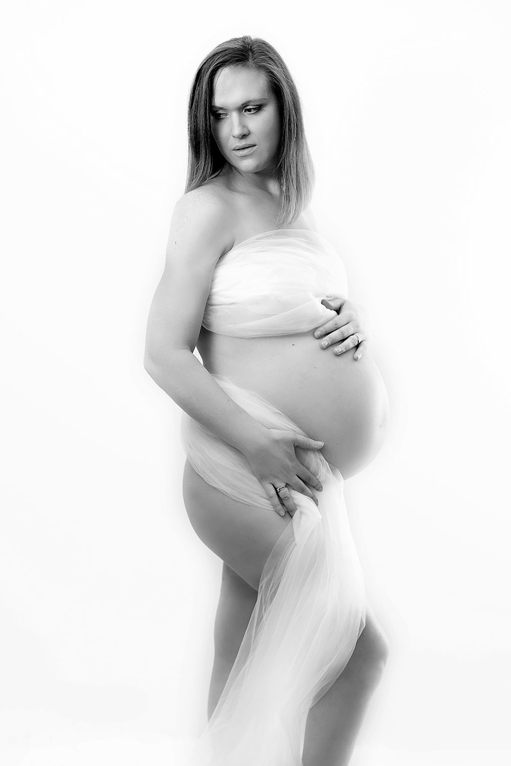 Black and white portrait of a pregnant woman draped in white sheer fabric holding her baby bump.