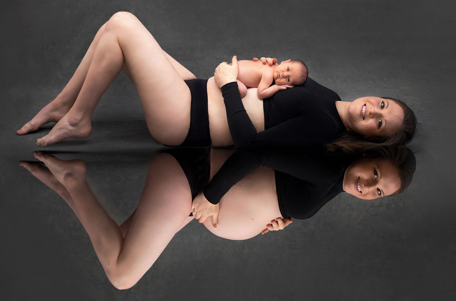Mother in black outfit lying on reflective floor holding newborn baby with mirror reflection below.