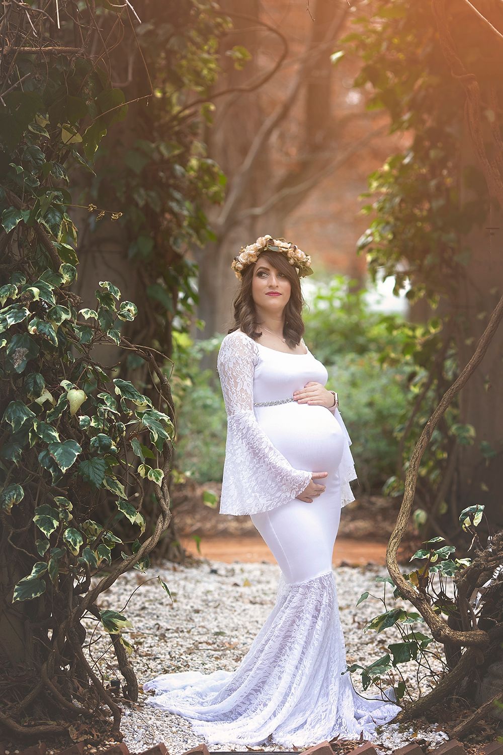 Pregnant woman in white lace gown with floral crown standing in a sunlit forest.