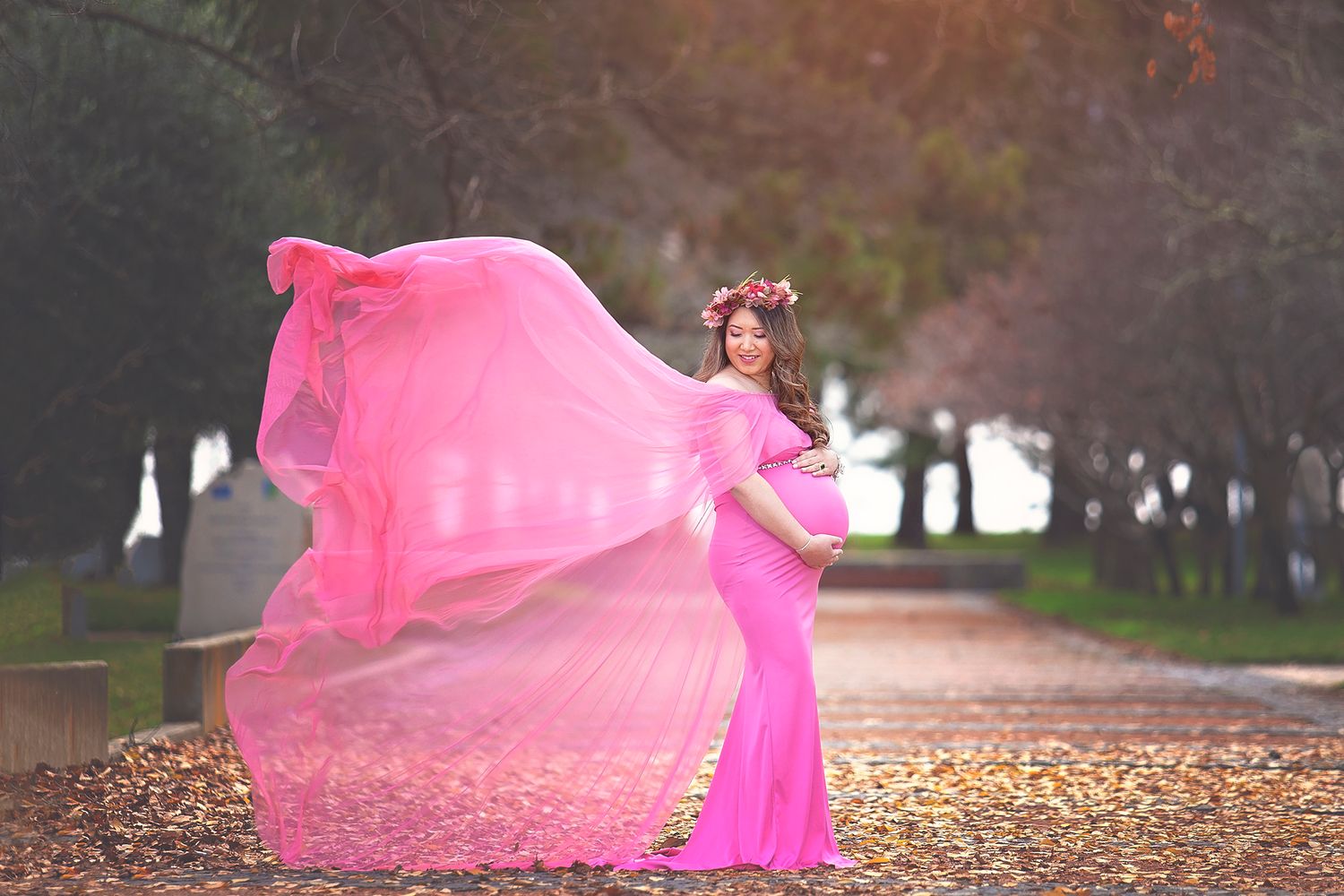 Pregnant woman in flowing pink dress with floral crown posing on an autumn tree-lined path.