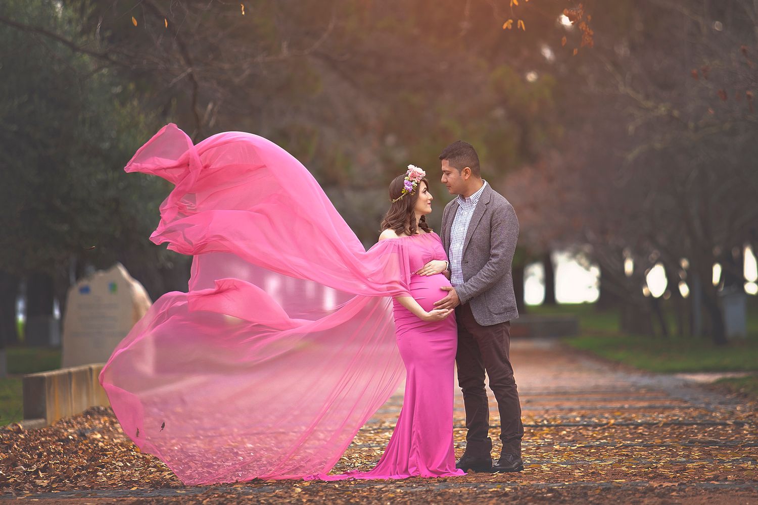Pregnant woman in flowing pink gown with partner in autumn park during maternity photoshoot.