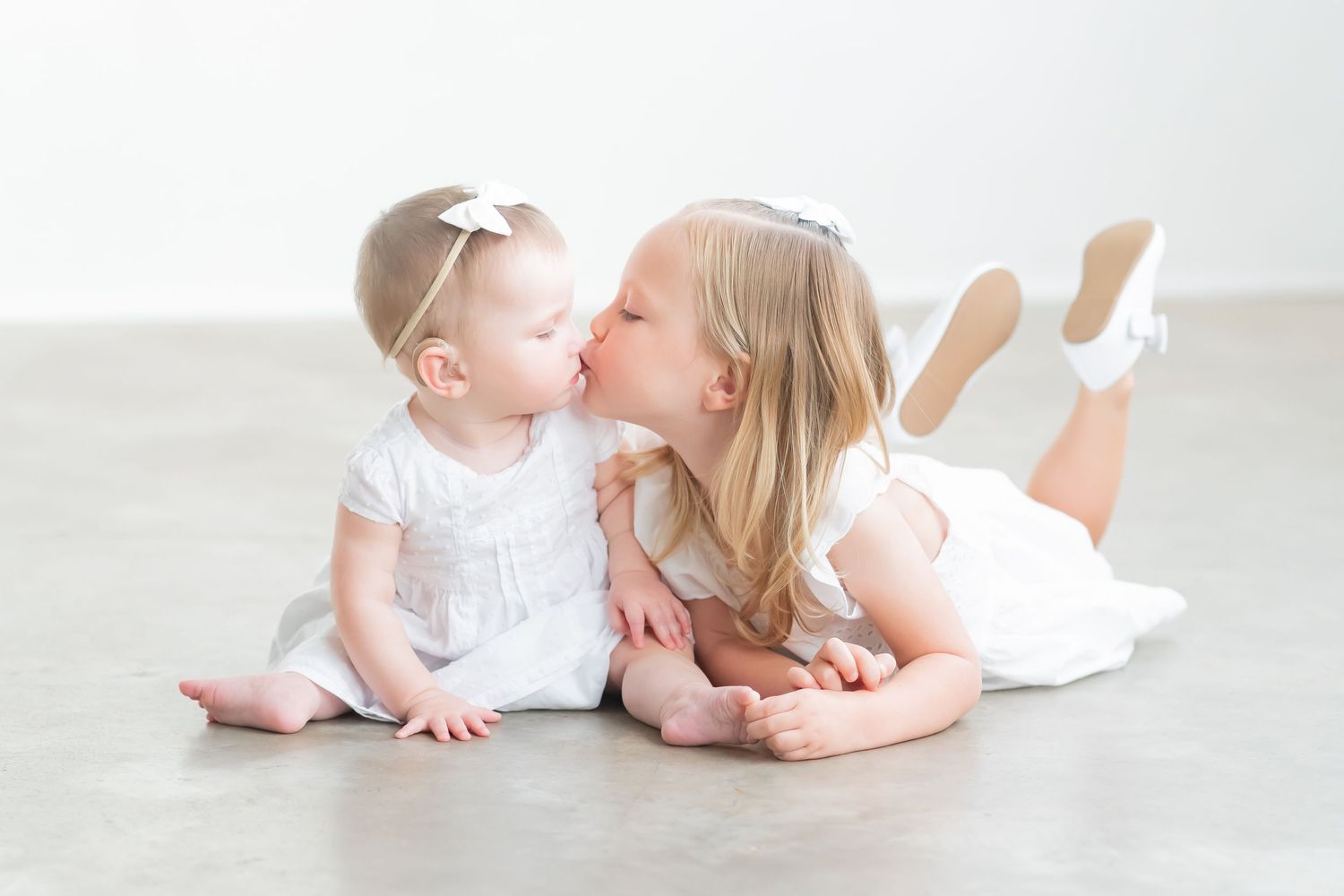 Two children in white dresses lying on floor sharing sweet moment together.