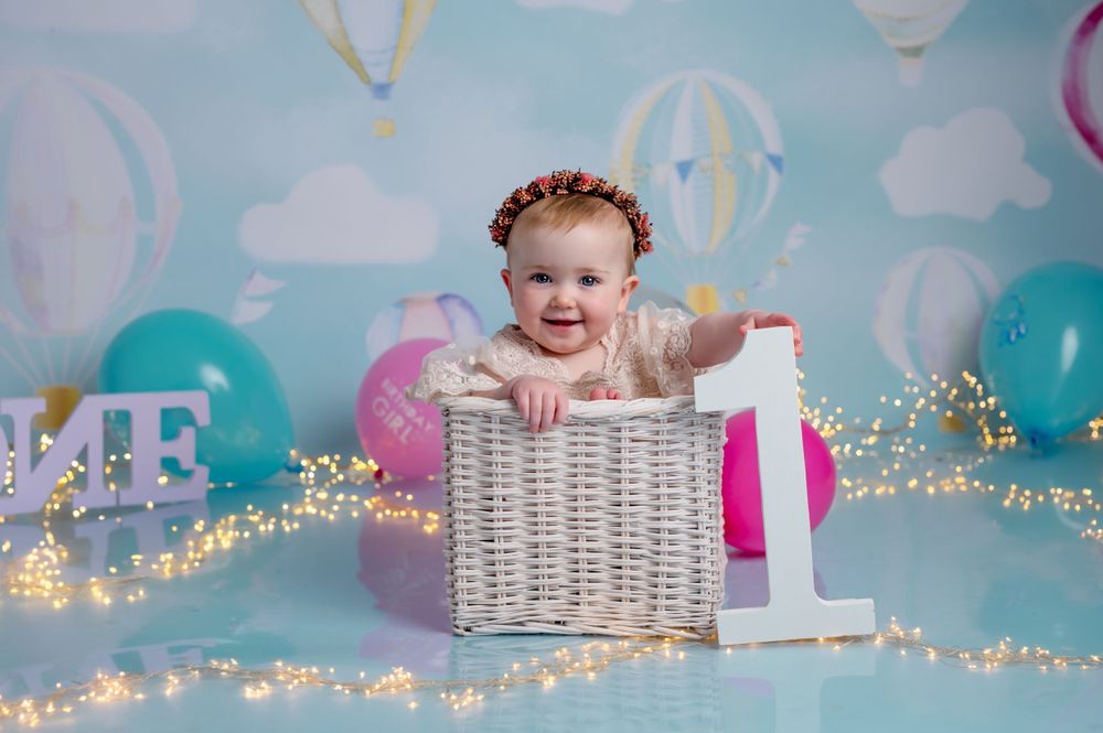 Whimsical first birthday photo with hot air balloon backdrop, sparkle lights, and decorative basket setup.