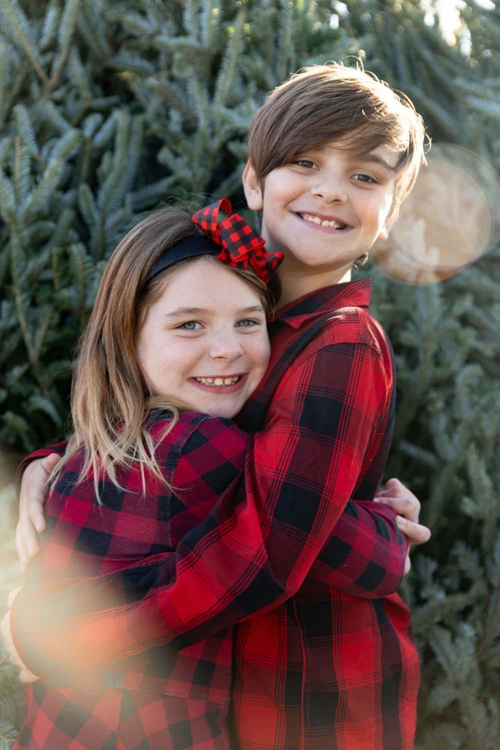 Two children in matching red and black buffalo plaid outfits embrace and smile while posing by evergreen trees.